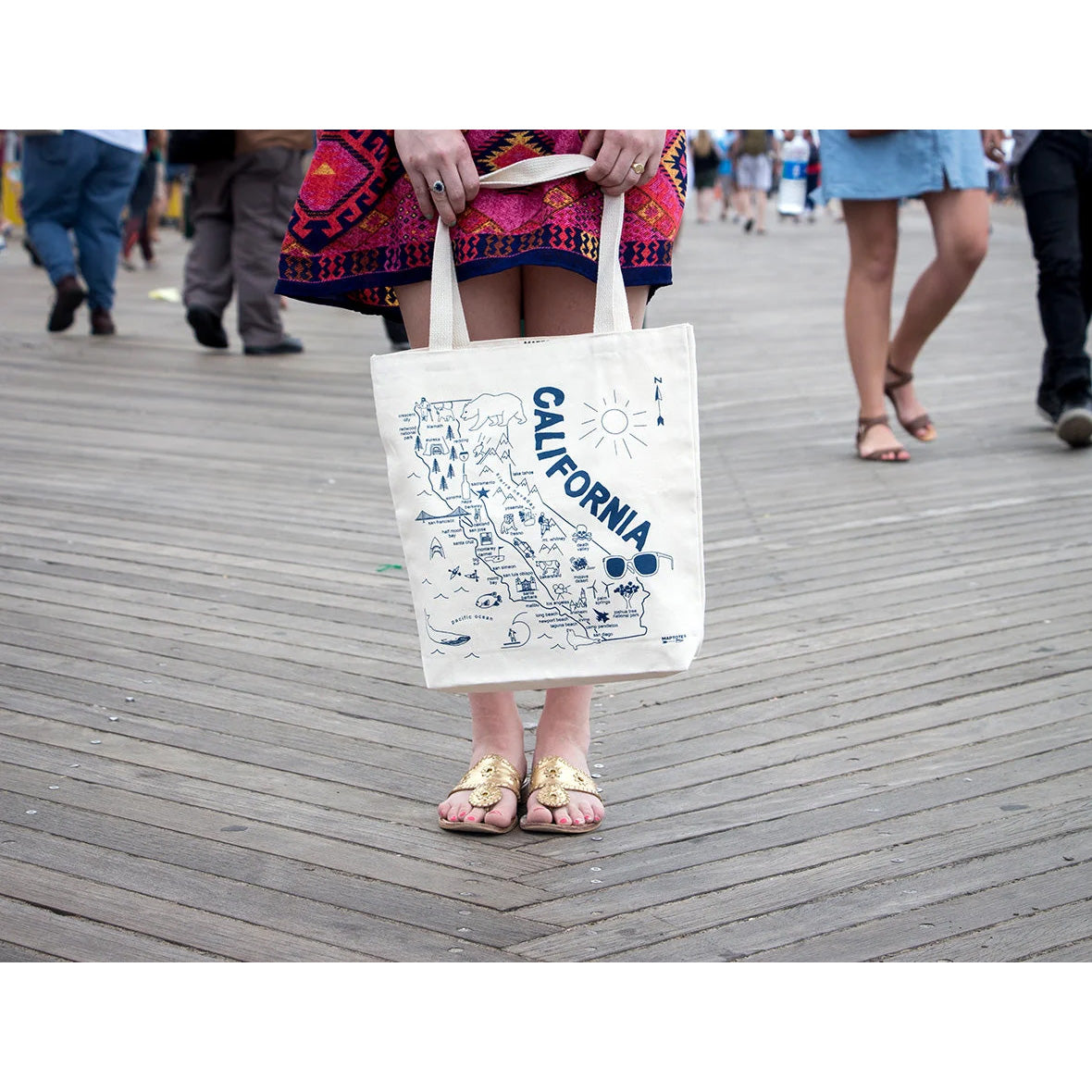 A person standing on a wooden boardwalk holds a cream canvas tote bag printed with a blue map illustration and the word 'CALIFORNIA.' The background shows blurred pedestrians in a real outdoor setting, making this a Lifestyle image.