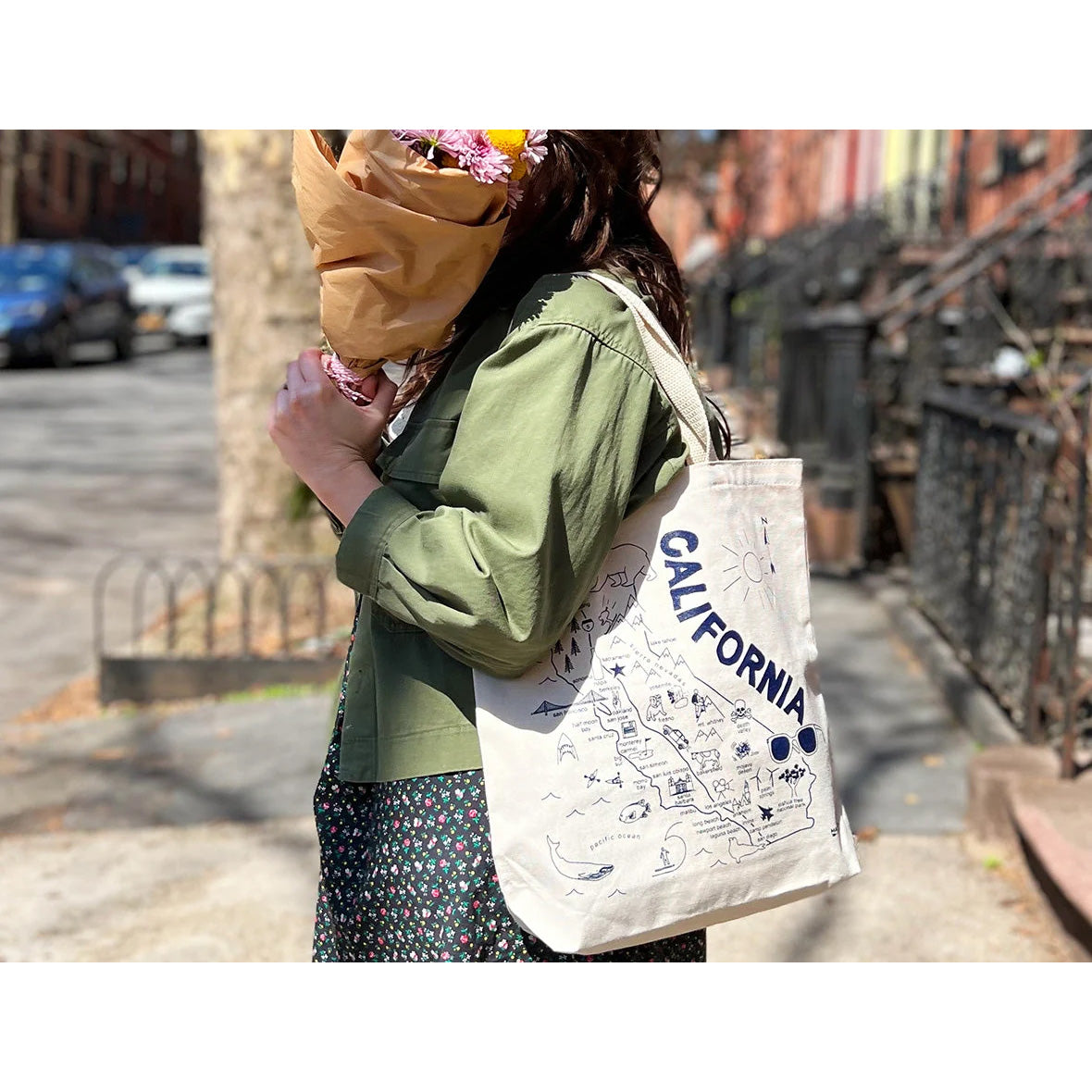 An off-white canvas tote bag featuring a blue line-art map of California with the word 'CALIFORNIA' written vertically, illustrated landmarks, and a sun graphic.
