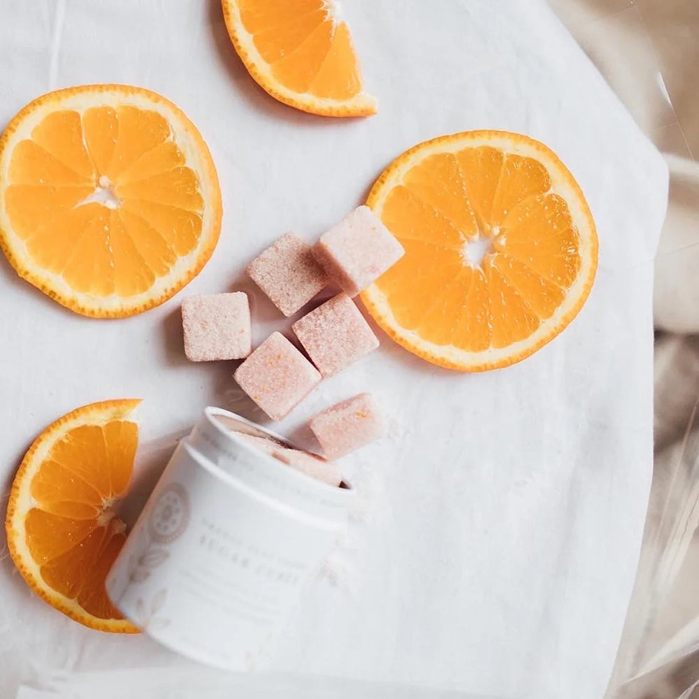 A tin container with orange zest cherry sugar cubes displayed in front of it, on a white background.