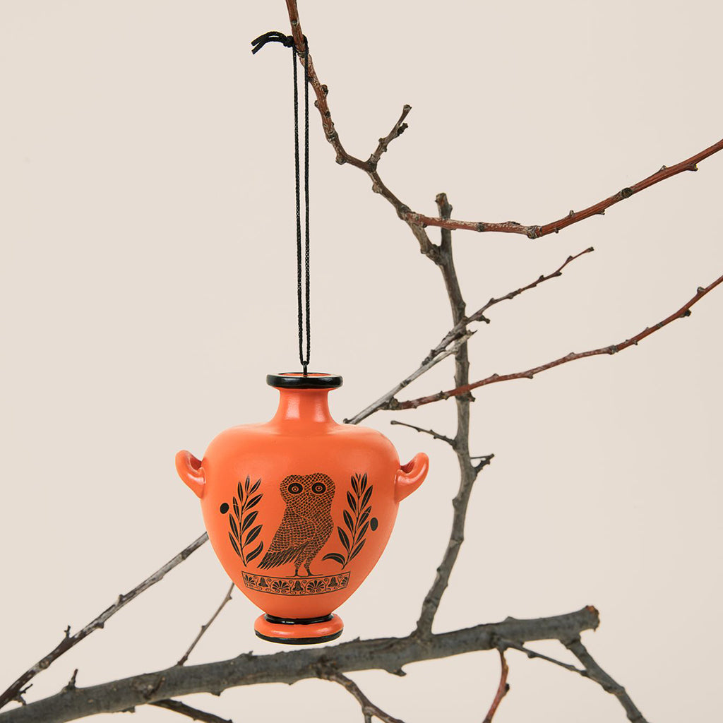 An orange ceramic Greek-style urn ornament hanging from a black string, decorated with a black owl and olive branch motif, displayed against a beige background on bare tree branches.