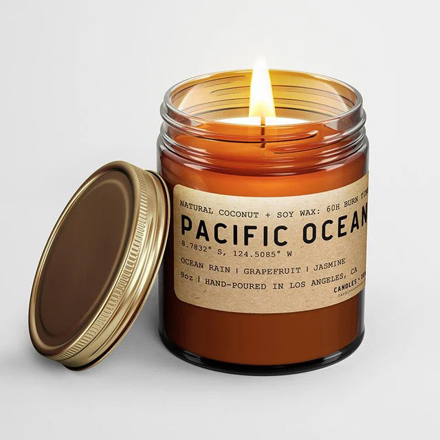A Pacific Ocean scented candle, with a label showing the name and fragrance notes of ocean rain, grapefruit, and jasmine. The candle is in an amber glass jar with a golden brass lid.