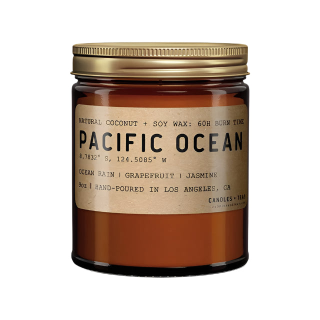 A Pacific Ocean scented candle, with a label showing the name and fragrance notes of ocean rain, grapefruit, and jasmine. The candle is in an amber glass jar with a golden brass lid.