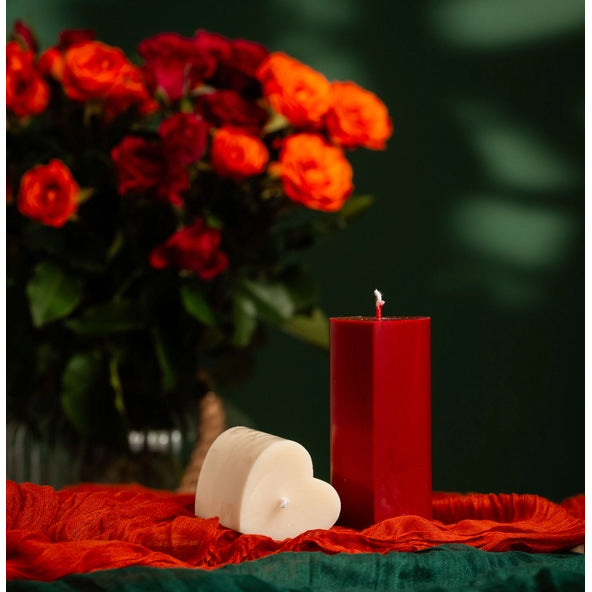Red candle and heart-shaped white candle on a red cloth with blurred red flowers in the background.