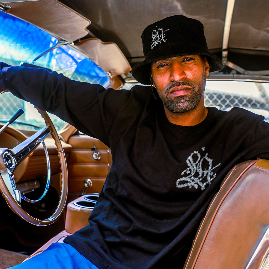 A man wearing a black bucket hat and a black long-sleeve t-shirt with a white graphic logo sits inside a vintage car with brown leather seats and a wooden steering wheel.