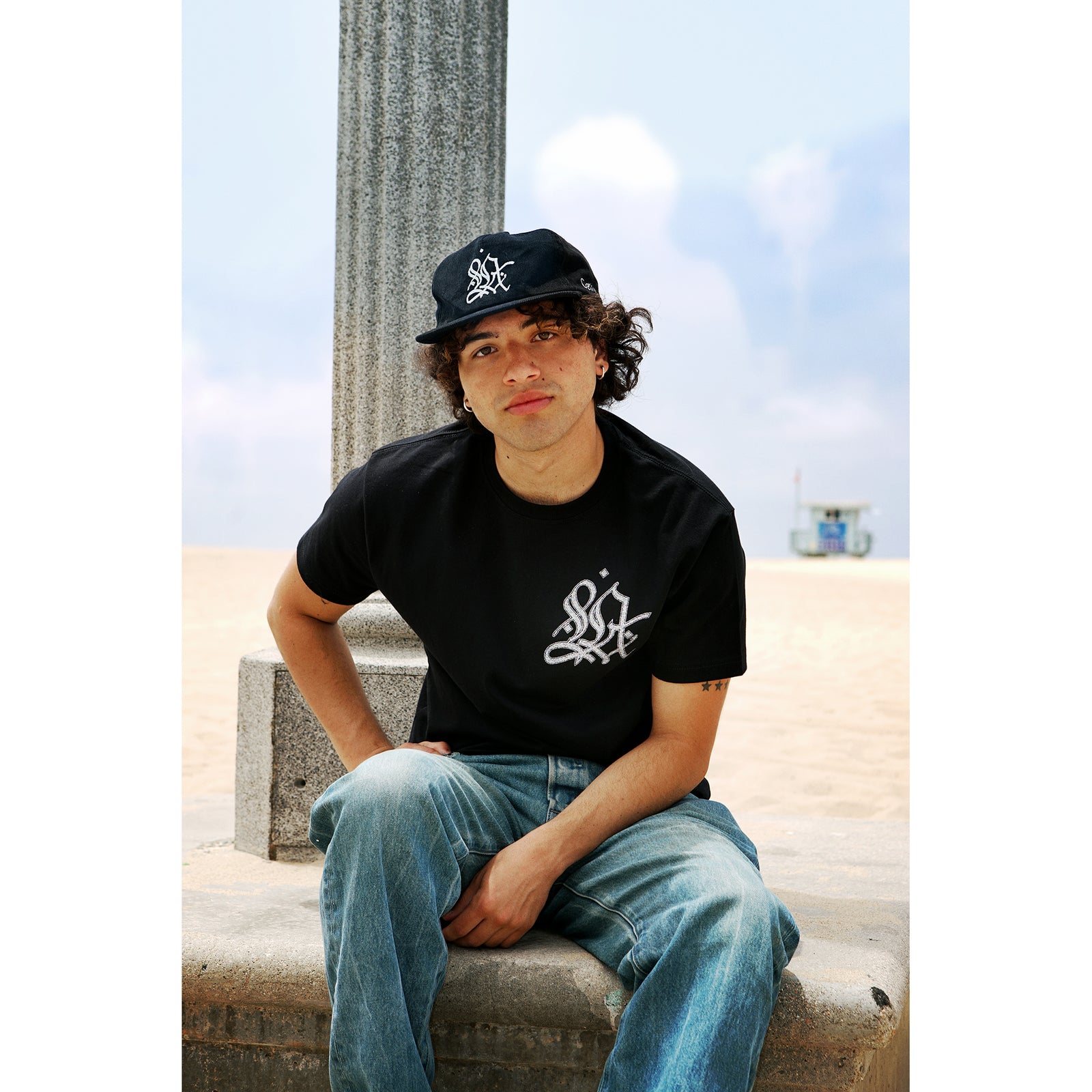 Person wearing a black t-shirt with a logo and blue jeans, sitting outdoors.