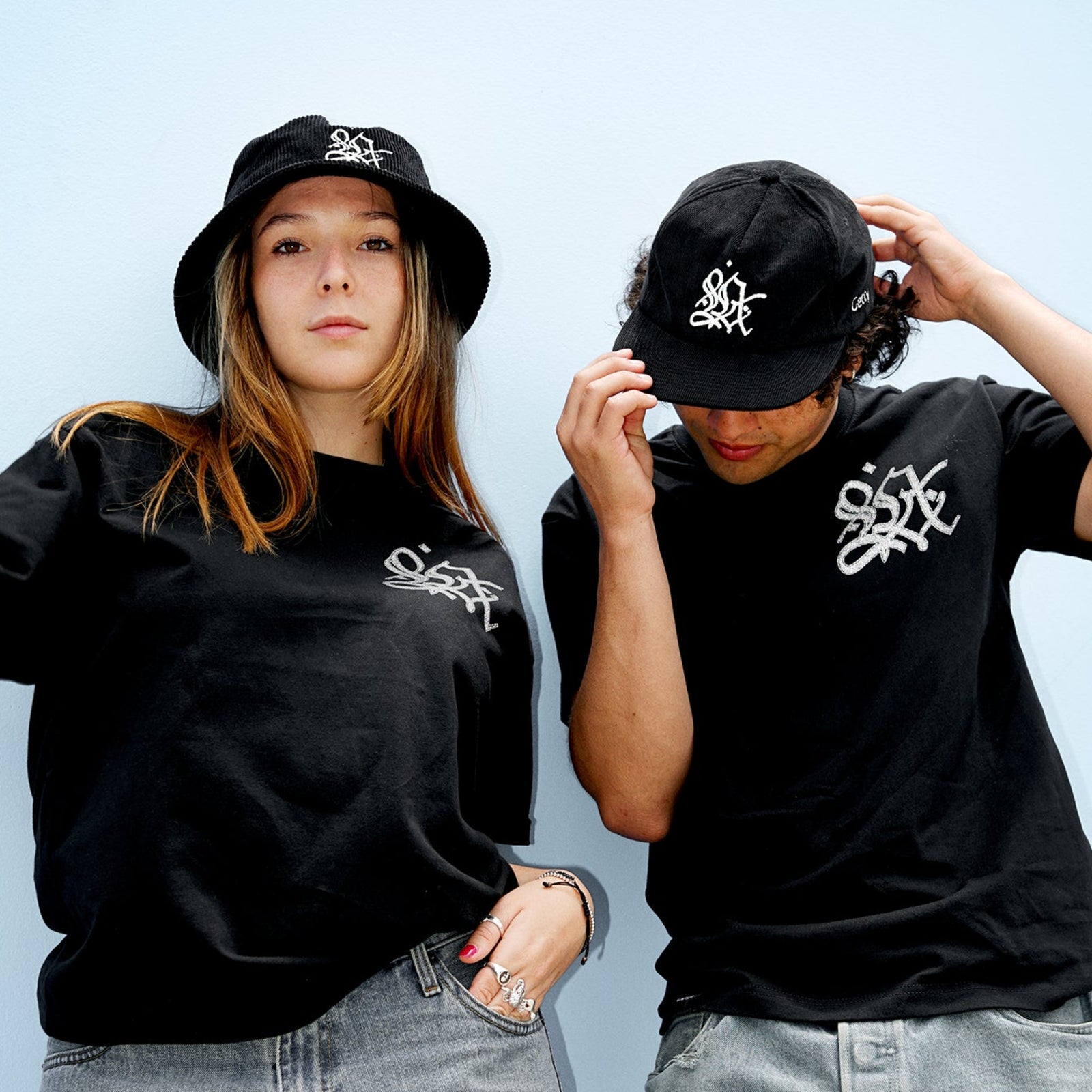 Two people wearing black t-shirts and hats with a logo, standing against a light blue background.