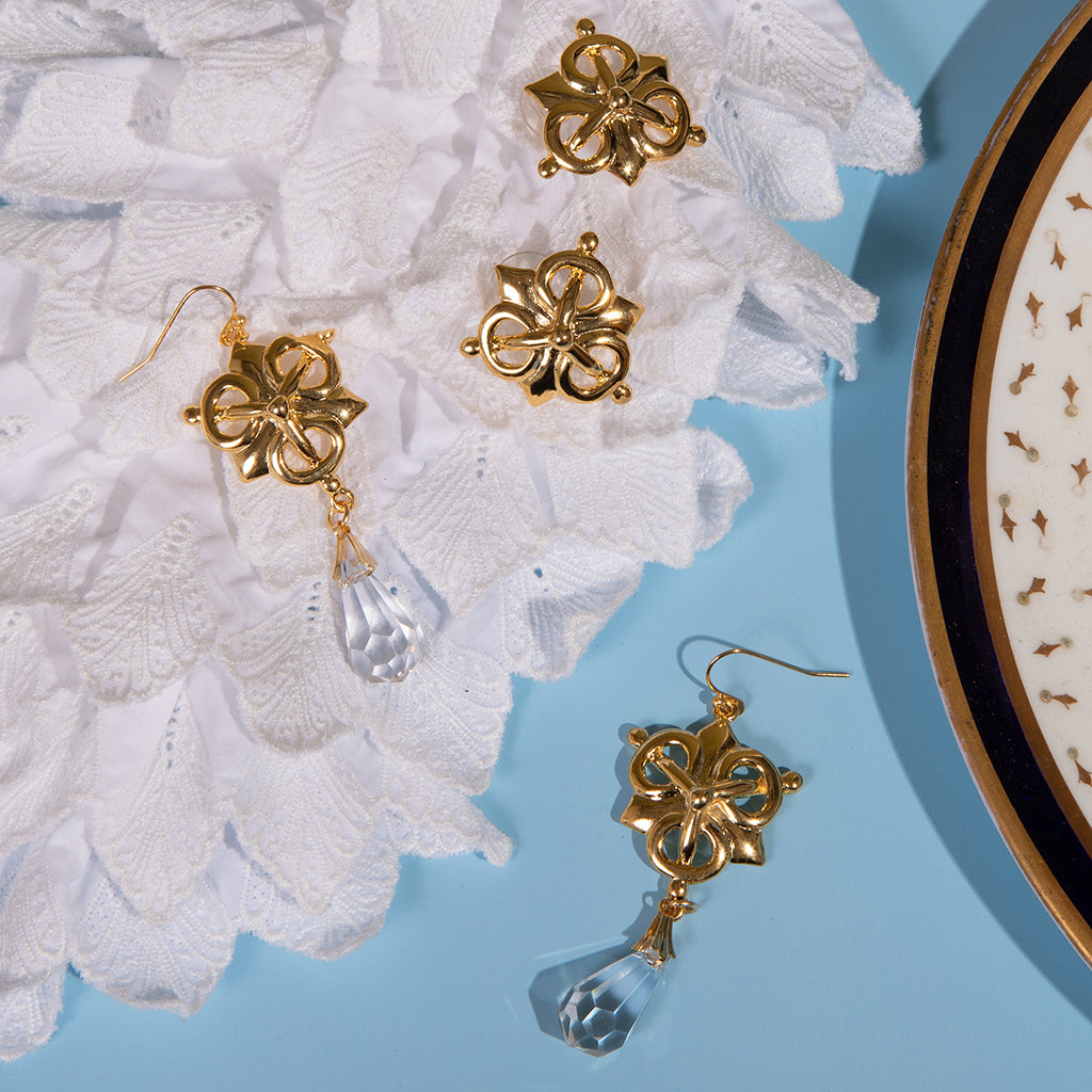 Gold-tone floral drop earrings with clear faceted crystal pendants, arranged on white textured fabric over a light blue background with a decorative plate edge visible on the right.