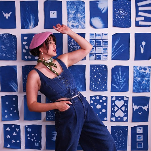 A woman posing in front of a wall covered in blue and white shibori dyeing samples, wearing a pink beret, green patterned neck scarf, and dark denim overalls.