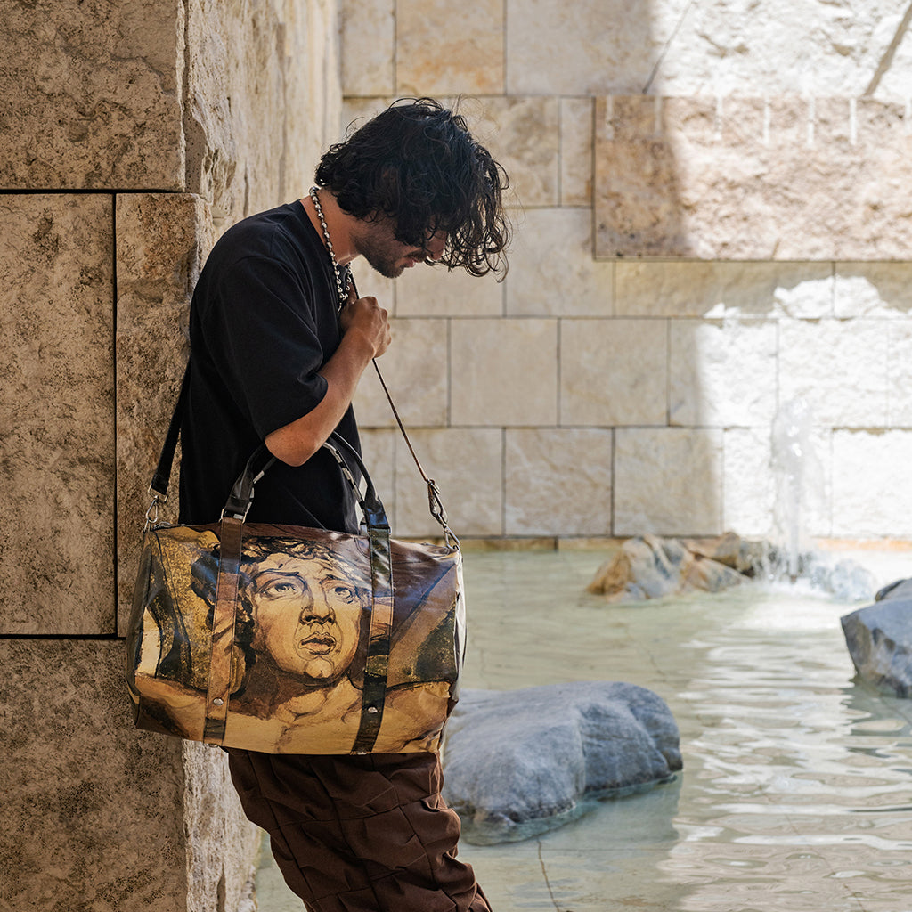 A man in a black t-shirt and brown pants stands by a stone wall next to a pool, carrying a large canvas tote bag printed with a classical bust portrait.