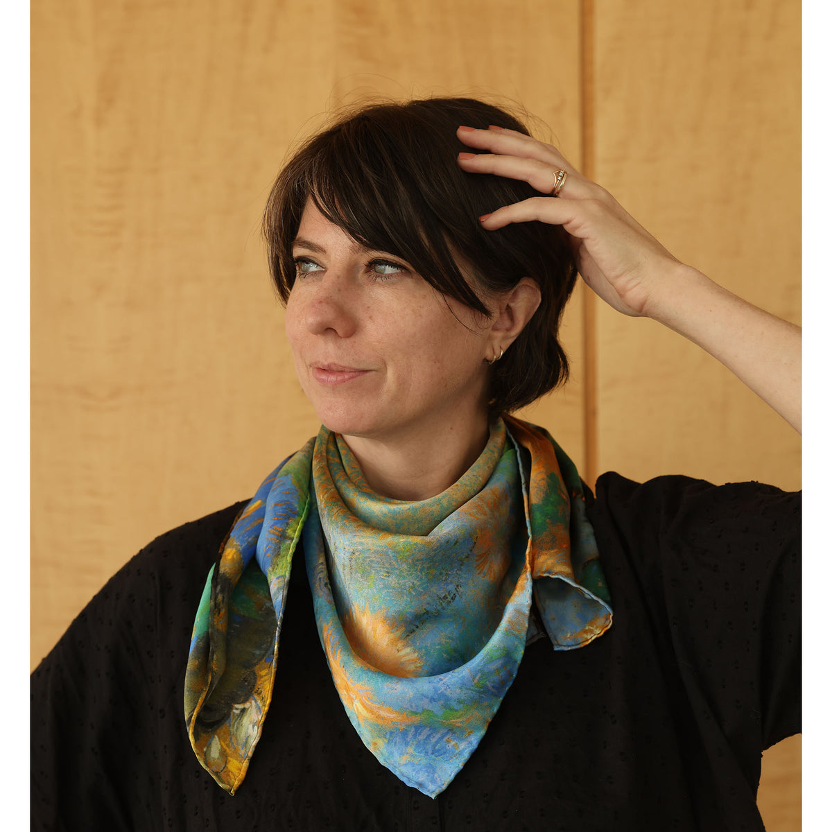 A woman with dark hair wearing a black top and a colorful patterned silk scarf in shades of blue, green, and gold, posed against a light wood-paneled background.