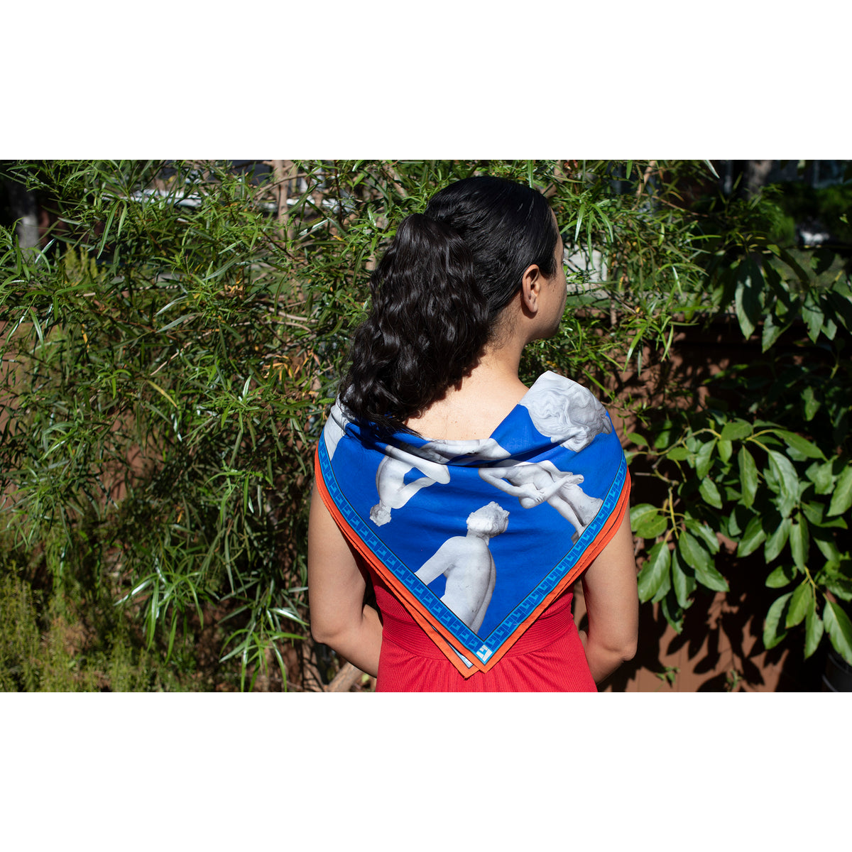 A woman viewed from behind wearing a red dress and a blue scarf with a white classical sculpture print and orange border, standing outdoors in front of green foliage.