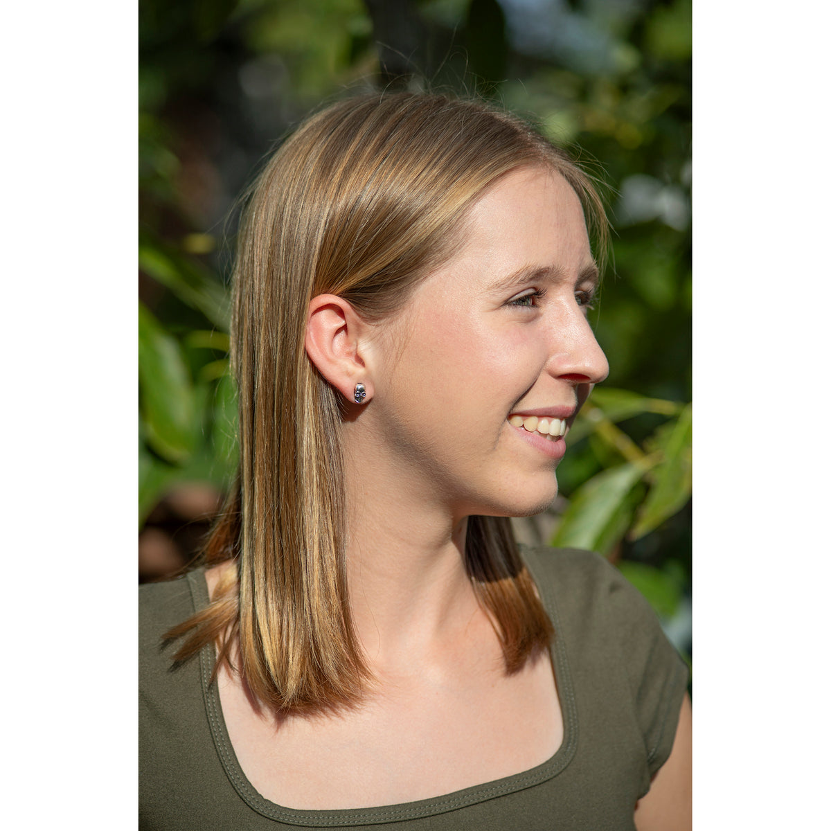 A woman with shoulder-length blonde hair is shown in profile outdoors, wearing a small silver stud earring shaped like a skull. She is wearing an olive green top against a blurred background of green foliage.