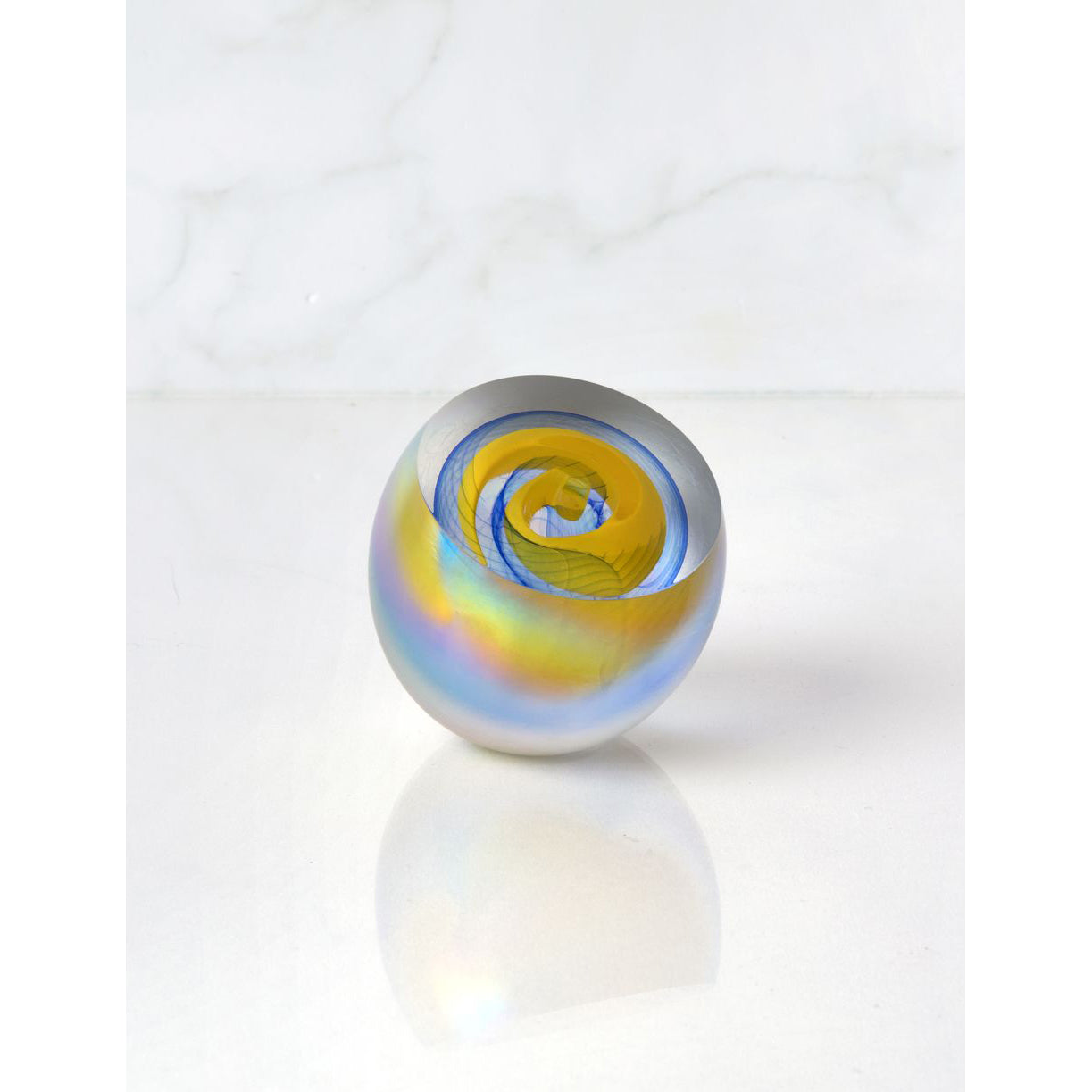 A round glass paperweight with blue and yellow swirl patterns and a mother of pearl appearance on the exterior.