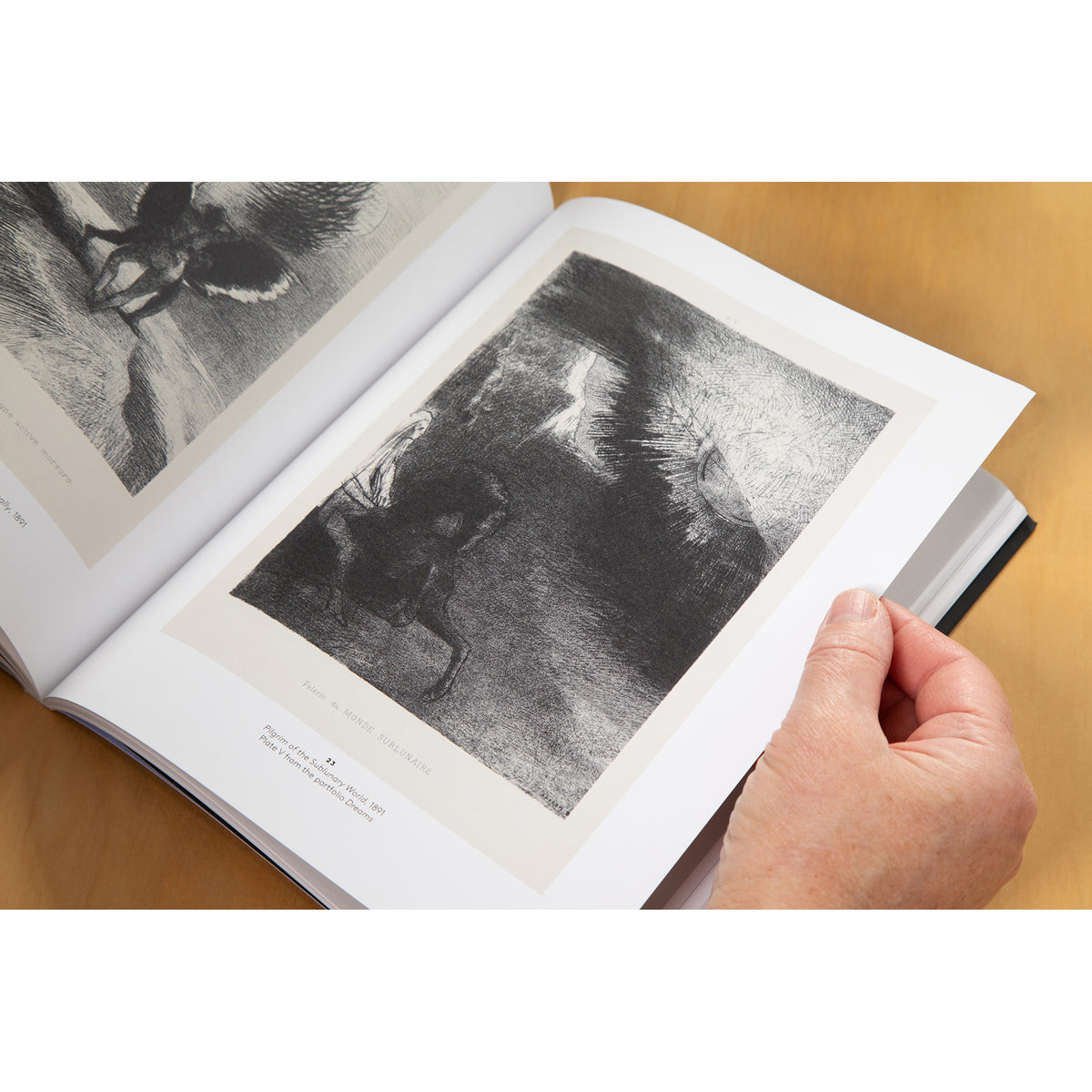 An open art book showing monochrome engraved artwork on white pages, photographed from above on a light wood surface with a hand holding the right page.