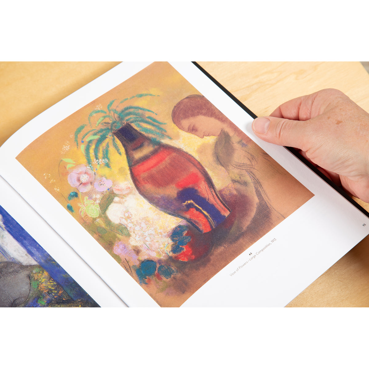 A hand holding an open art book page showing a colorful still-life painting of a red bottle with flowers and green leaves on a warm beige background.