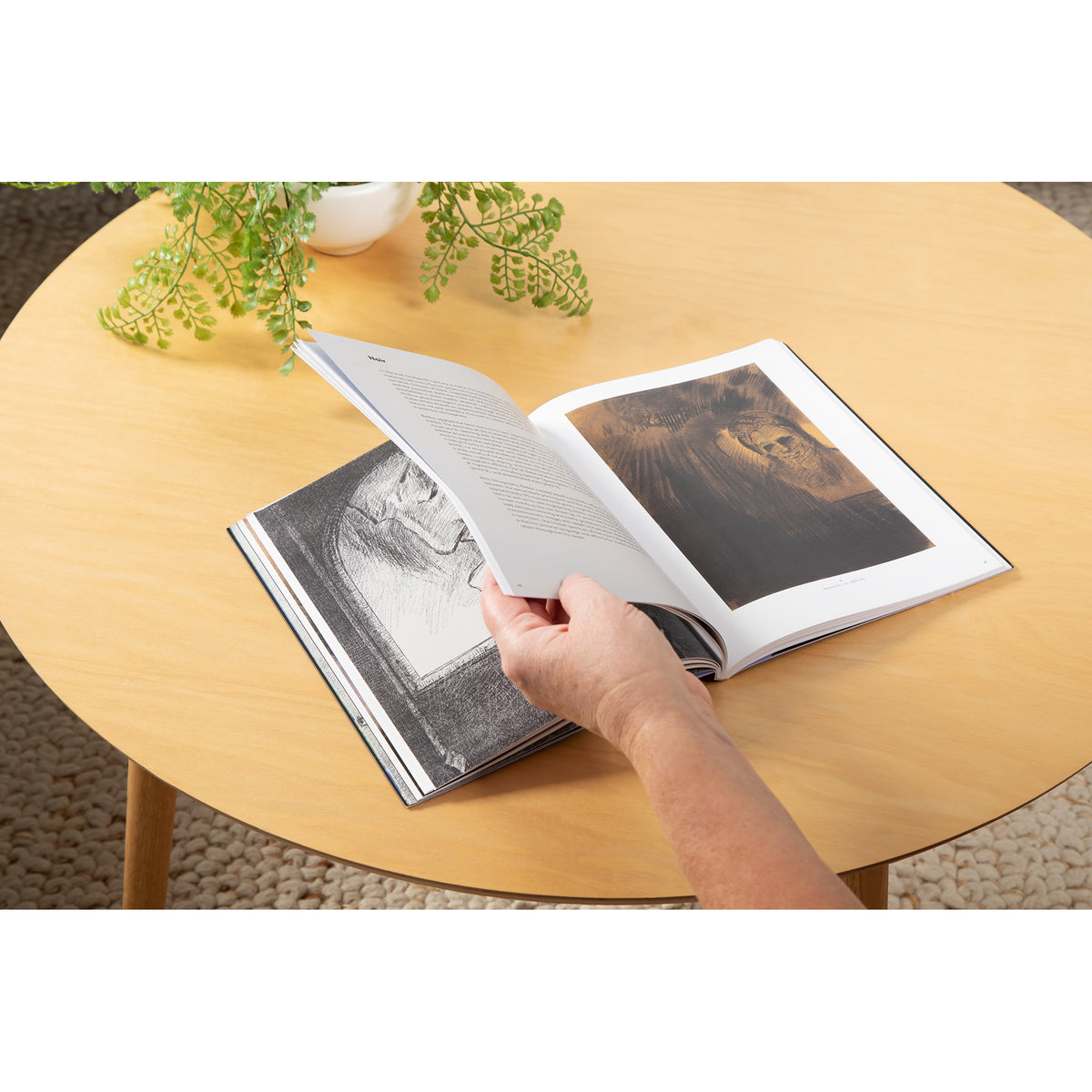 A hand holding an open page of a large black-and-white art book on a round light wood table, with a plant in the background.