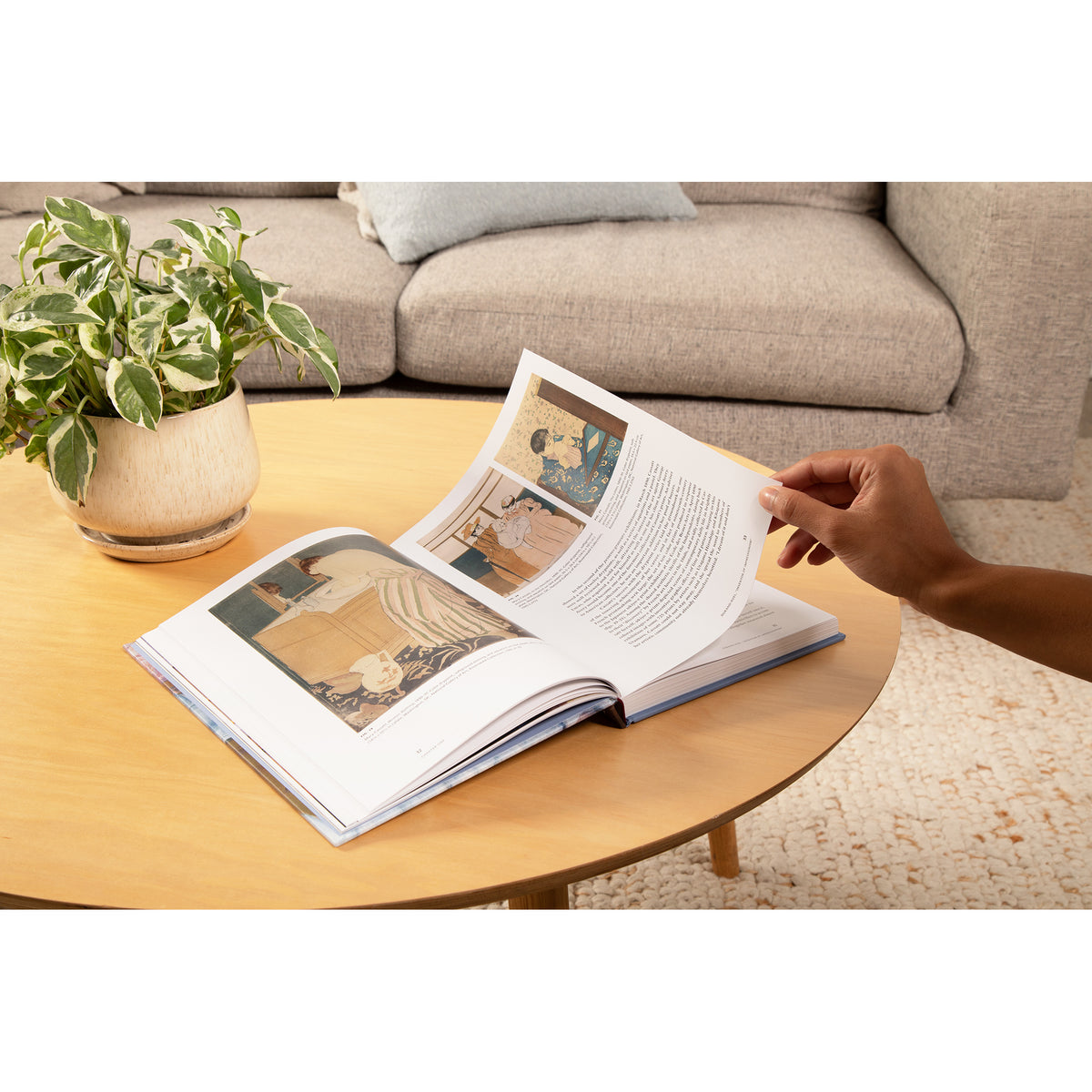 An open book lies on a round wooden coffee table in a living room, with a person's hand turning a page. A potted plant and a beige sofa are visible in the background.