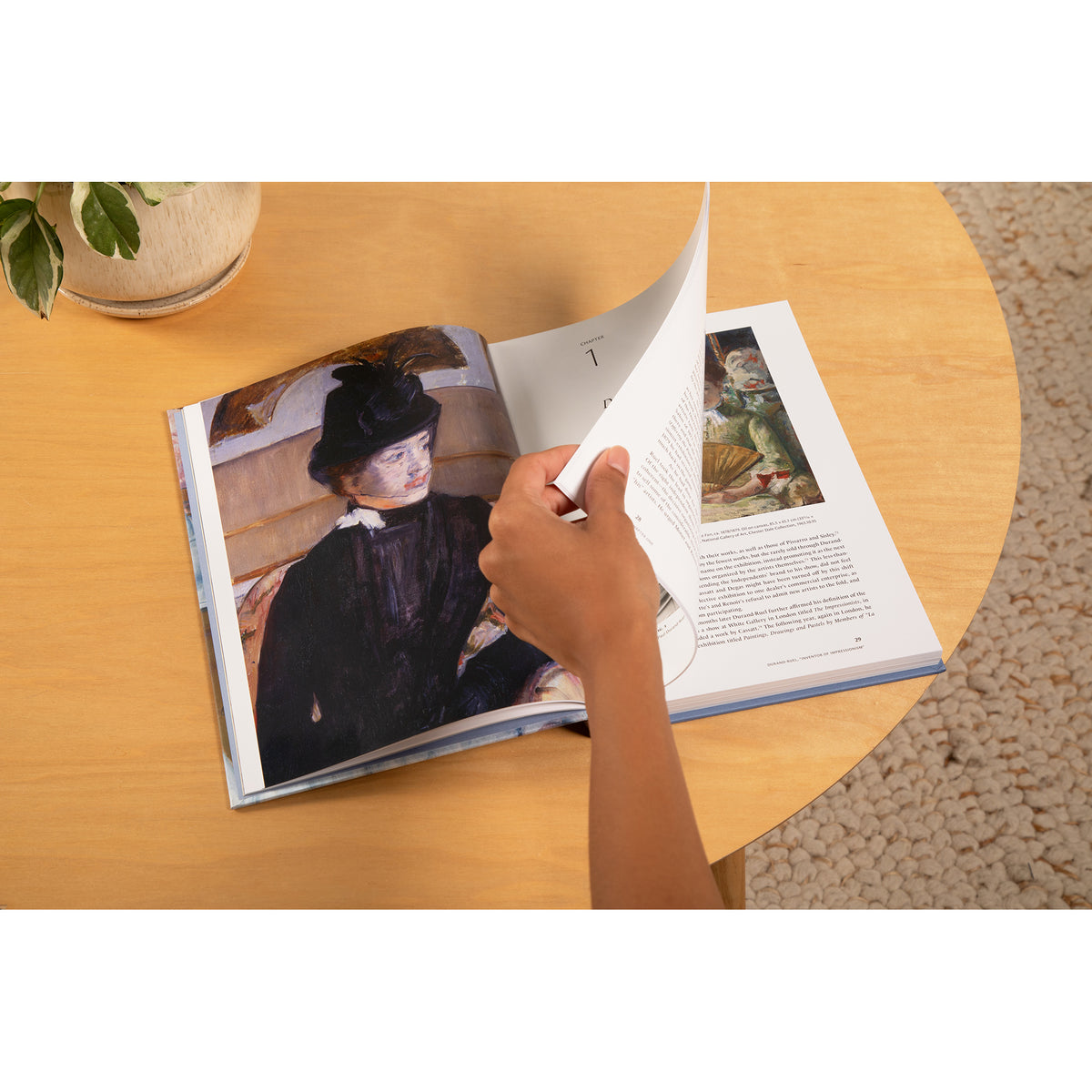 A hand turning a page of an open hardcover book on a light wood table, with a painting and text visible on the spread.