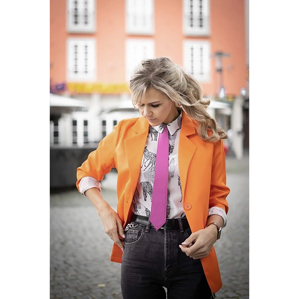 Woman wearing an orange coat with a pink tie and black pants on a street.