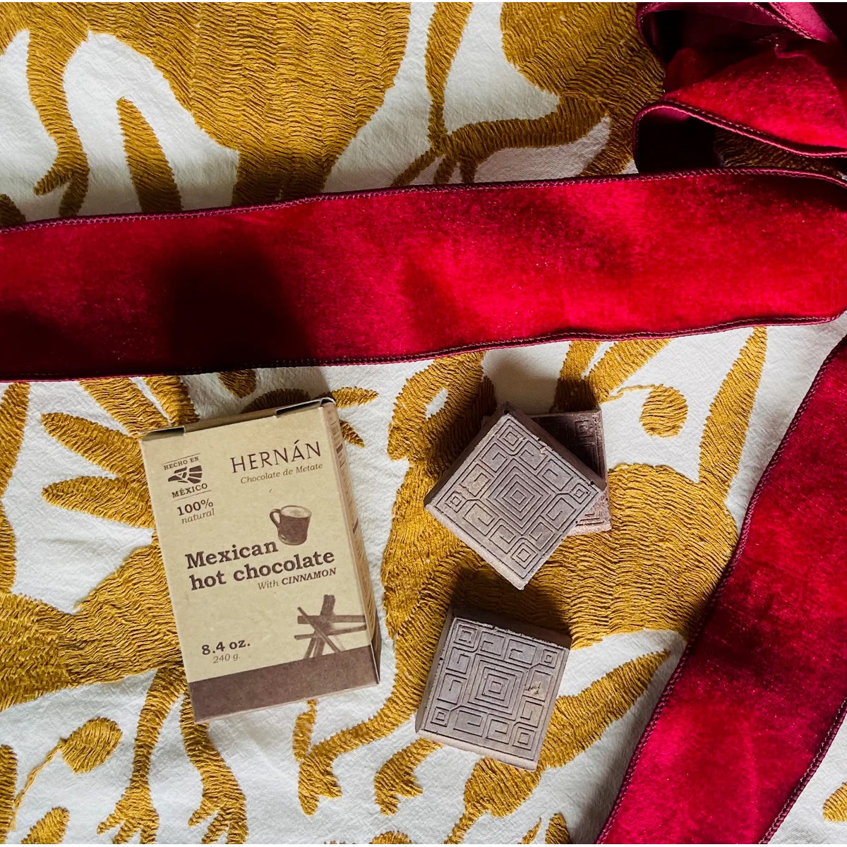 A small box of Hernán Mexican hot chocolate with chamomile alongside two square chocolate pieces, arranged on a cream and gold patterned fabric with a red velvet ribbon.