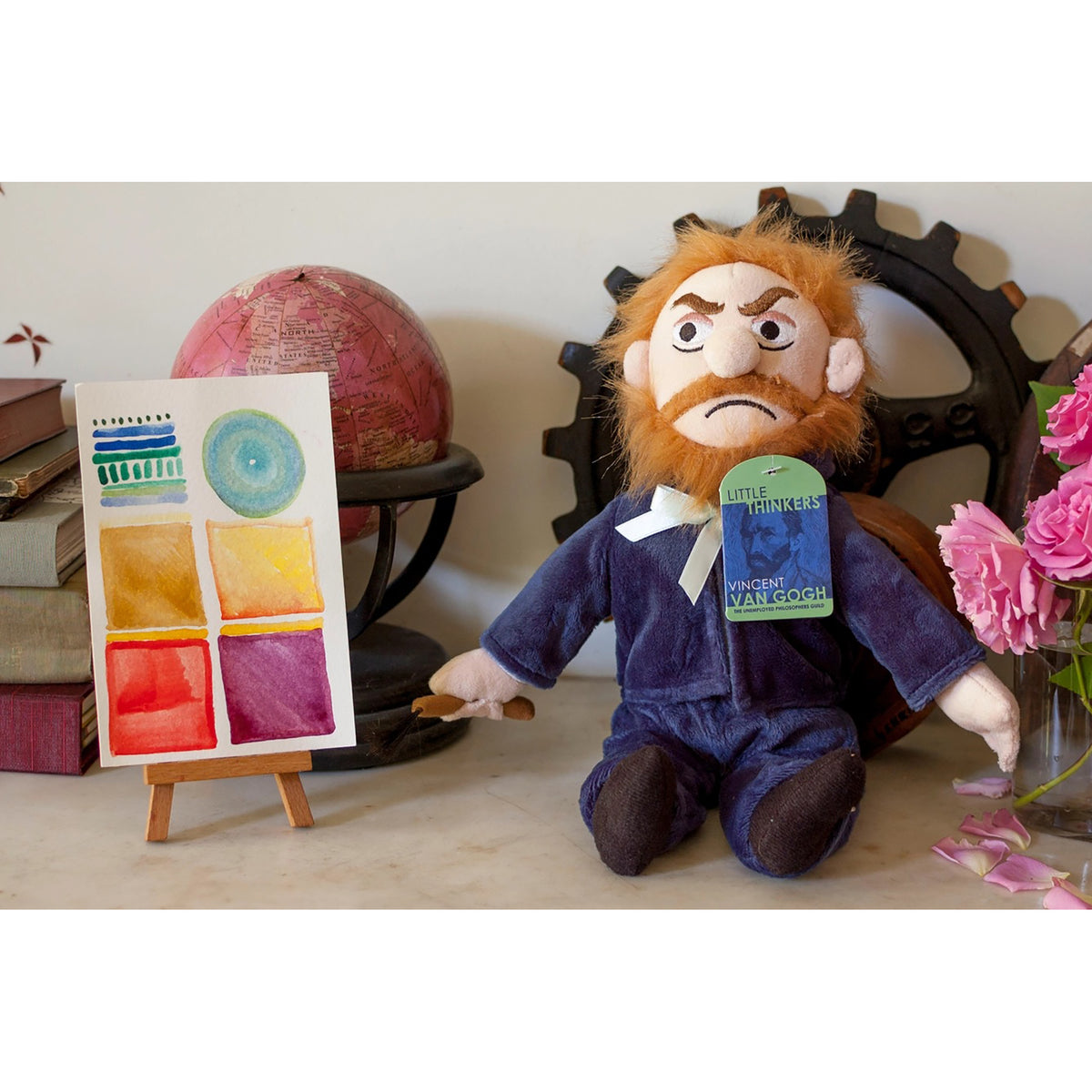 A plush doll styled as Vincent van Gogh with orange hair and beard, wearing a dark blue suit and holding a paintbrush, seated next to a small painted color swatch canvas on a tabletop with books, a globe, gears, and pink flowers in the background.