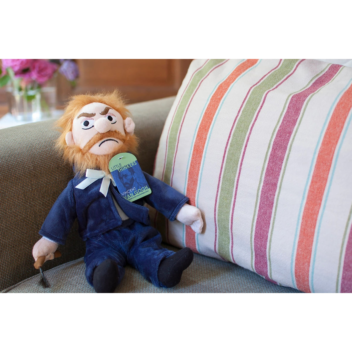 A plush doll that resembles Vincent van Gogh with a reddish-orange beard, wearing a blue suit and holding a cane, resting on a gray upholstered chair next to a striped cushion.