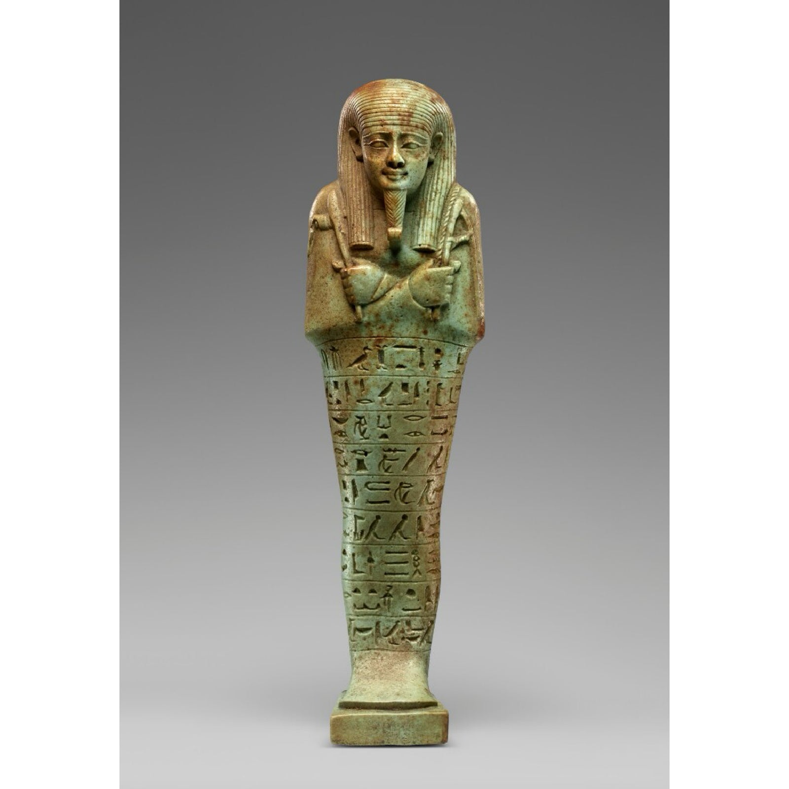 An enamel pin shaped like an Egyptian ushabti funerary figurine, in green color, standing on a rectangular base with a hieroglyphic inscription.