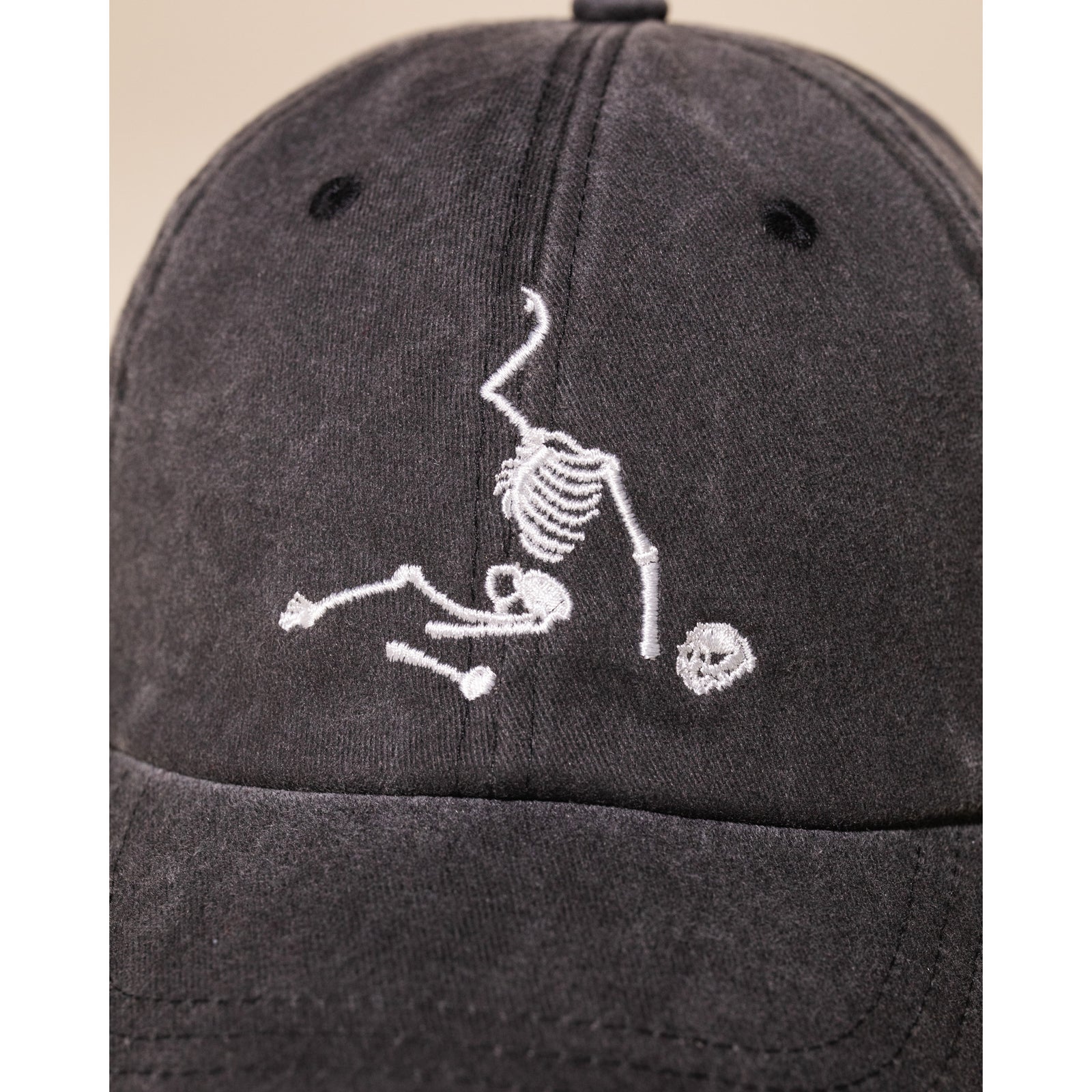 A dark gray washed baseball cap featuring a white embroidered graphic of a skeleton holding a soccer ball on the front, set against a plain white background.