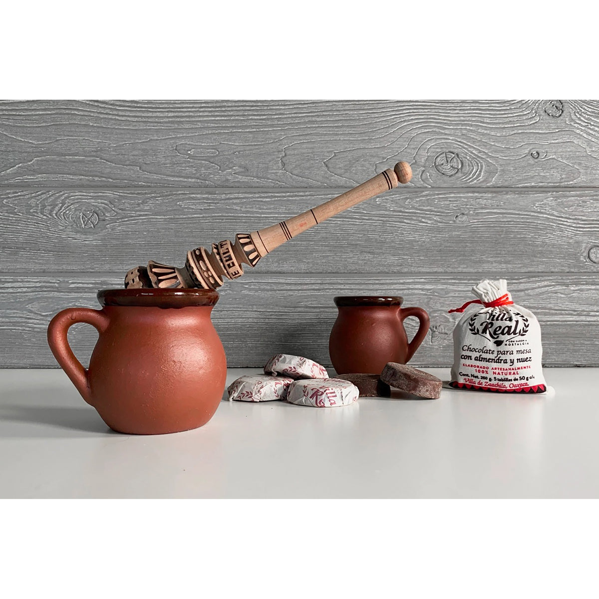 A rustic terracotta clay mug set with a wooden spoon, two small clay cups, several unwrapped dark chocolates, and a bag of 'hot chocolate' on a white surface against a gray wood-plank background.