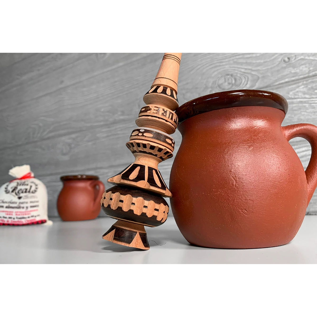 A terracotta-colored ceramic pitcher with a dark brown rim and handle, standing next to a tall, patterned wooden or clay tower vessel on a light gray surface. A small white bag is blurred in the background against a gray wood-textured wall.