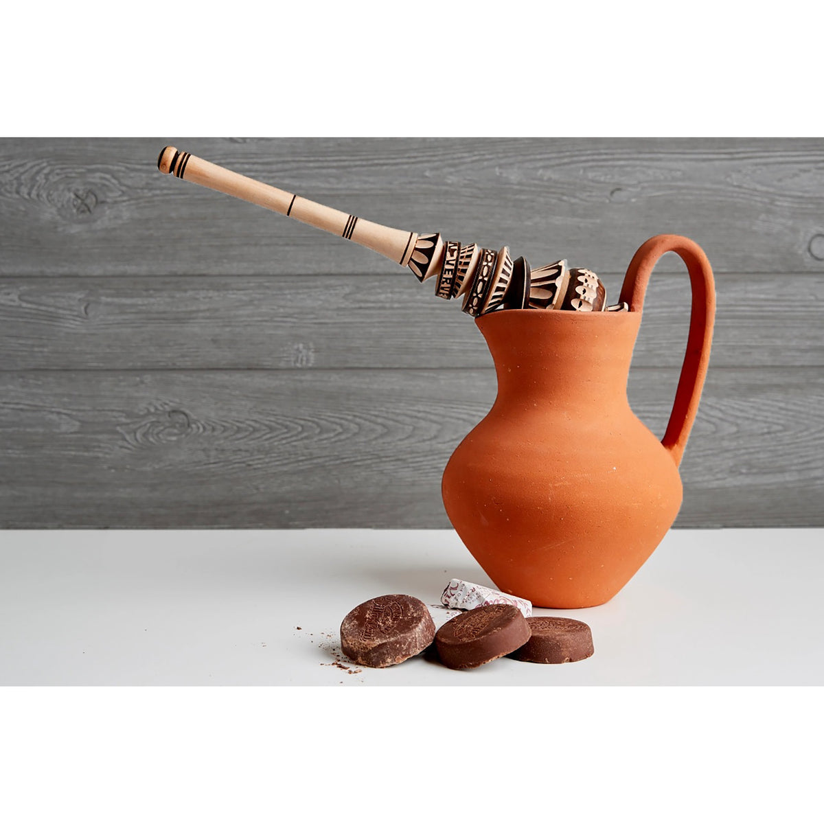 An orange terracotta pitcher with a long handle and a carved wooden spoon resting inside, set on a white surface against a gray wood-plank background. Three chocolate pieces are placed in front of the pitcher.