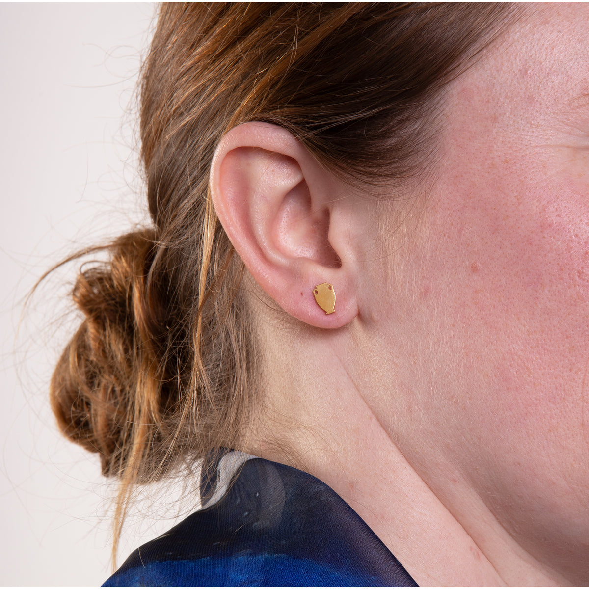 Close-up of a person wearing a gold vessel-shaped earring on a plain background