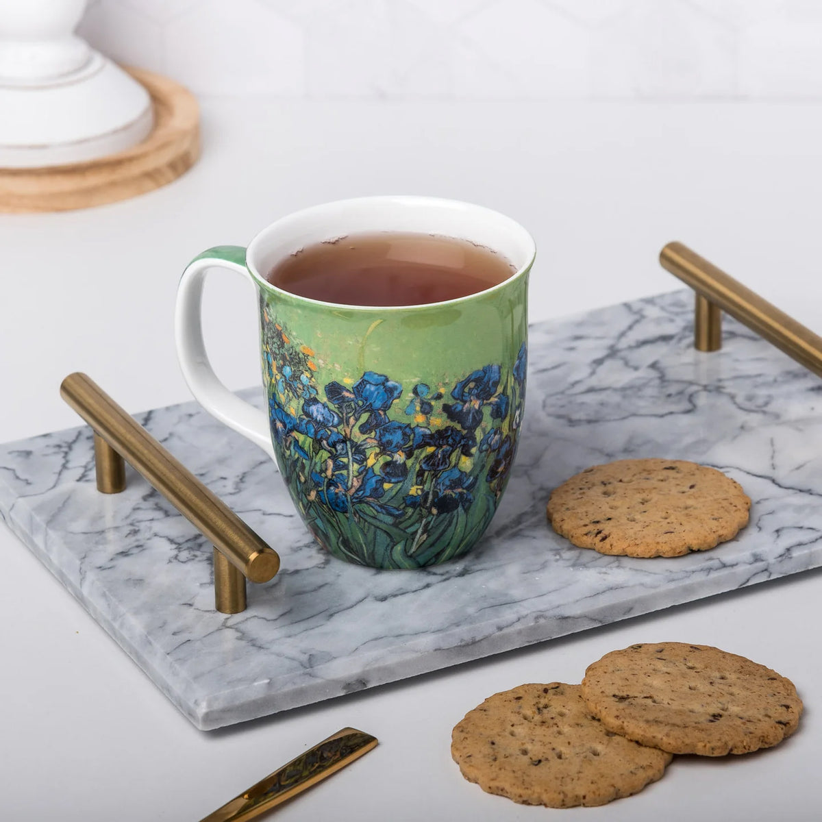 A ceramic mug with a blue and green floral design, filled with tea, sits on a marble serving tray with gold-toned handles. Two cookies are placed beside the tray on a white surface.