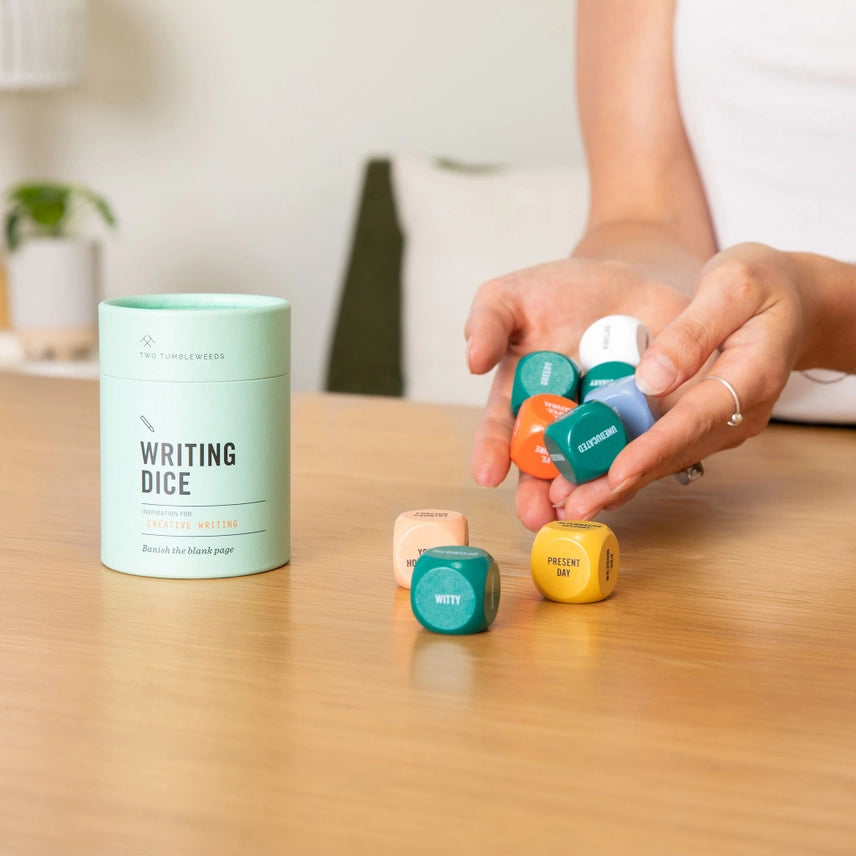 Person holding colorful dice next to a 'Writing Dice' container on a wooden table.
