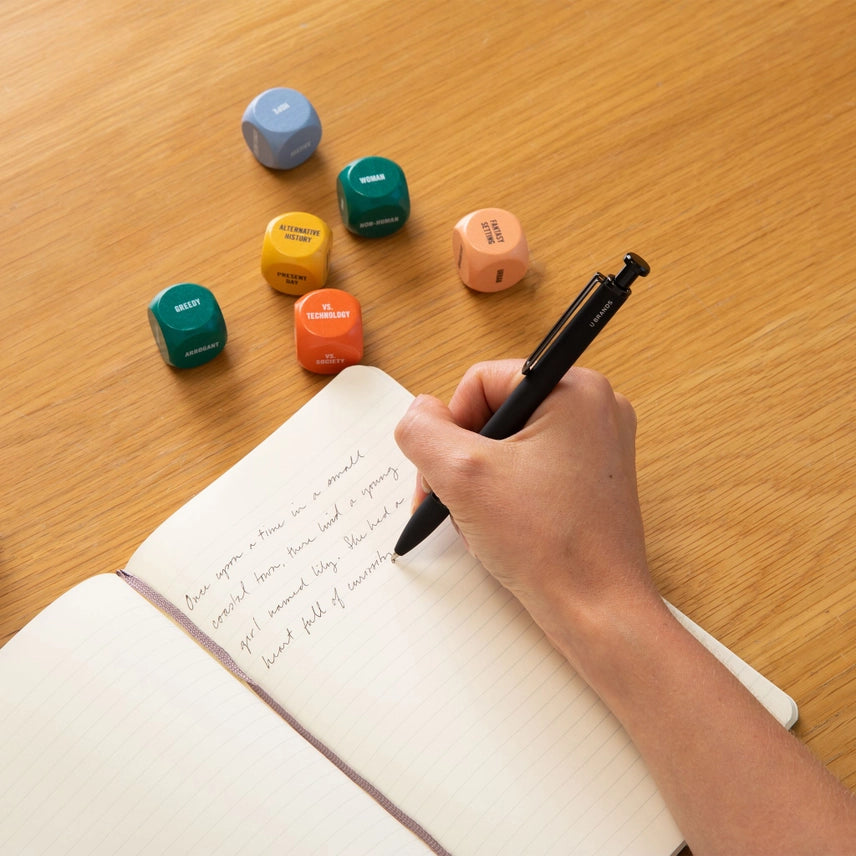Hand writing in a notebook with colorful dice on a wooden surface