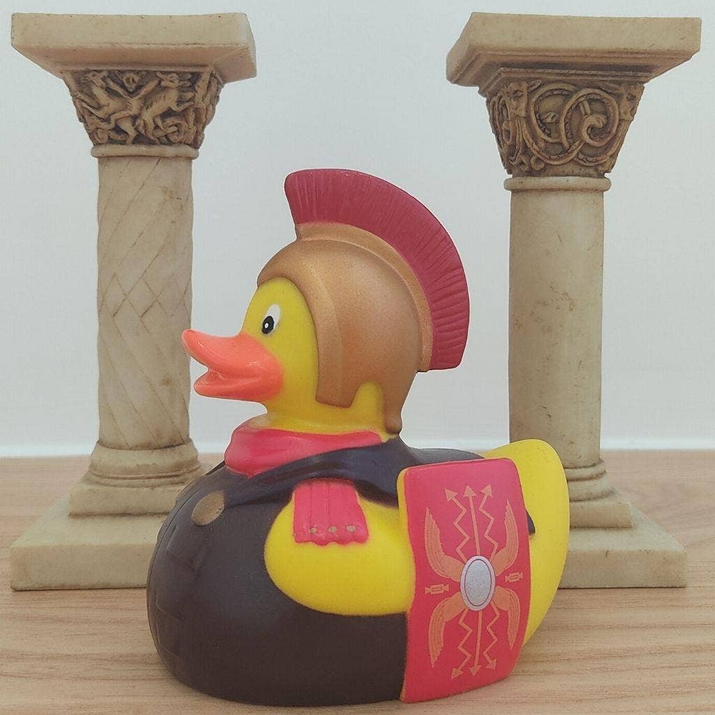 A rubber duck styled as a Roman soldier, featuring a yellow body, brown helmet, red crest, pink scarf, and shield, set against two decorative stone columns on a wooden surface.