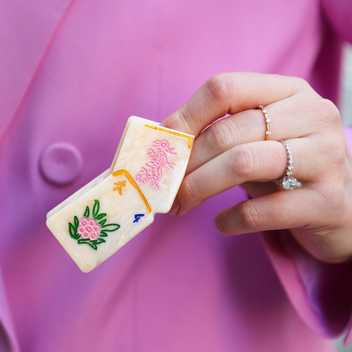 A close-up of a hand holding a small white hair clip decorated with embroidered fish and floral motifs in pink, green, yellow, and blue, worn with a pink blazer and rings.