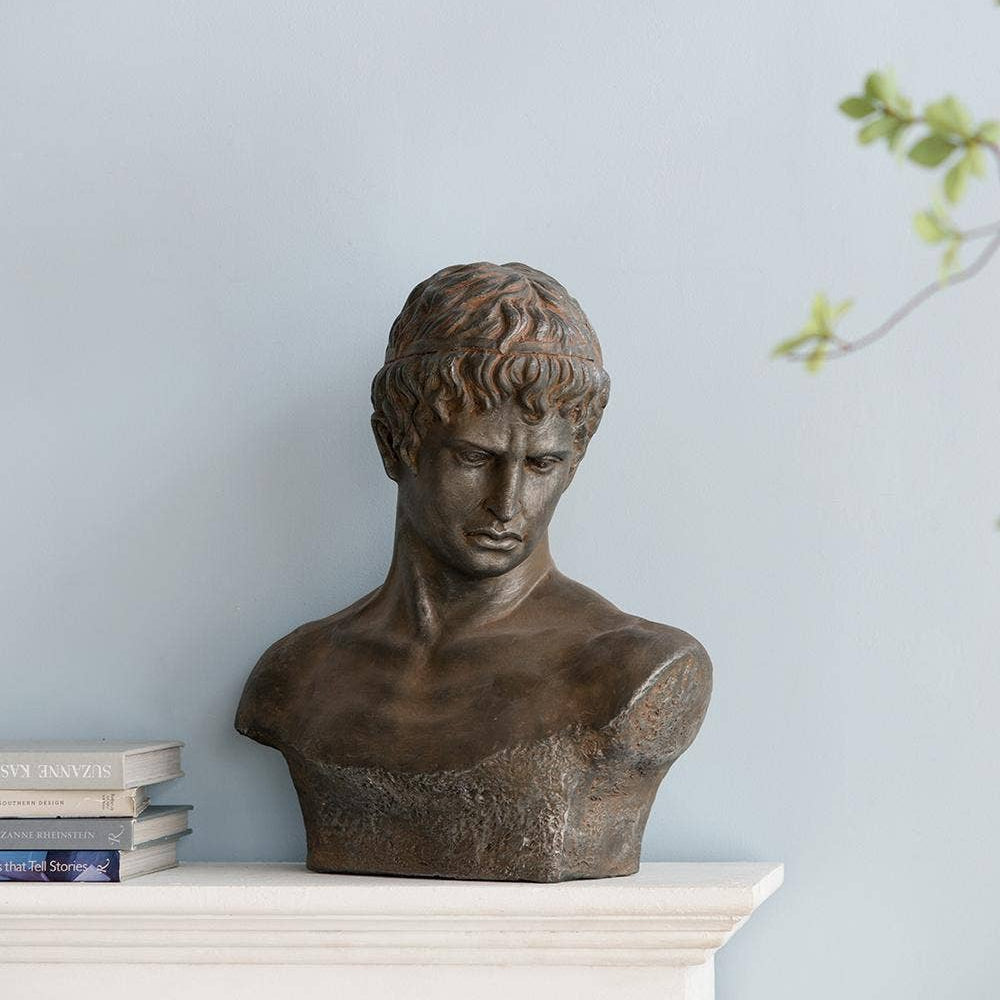 A bronze-finished classical male bust sculpture displayed on a white mantelpiece against a pale blue wall, with a small stack of books to the left and a leafy branch entering from the upper right.