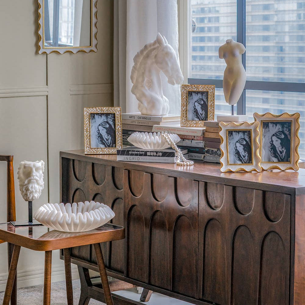 Decorative setup with sculptures, framed pictures, and a wooden console table in a room with large windows.