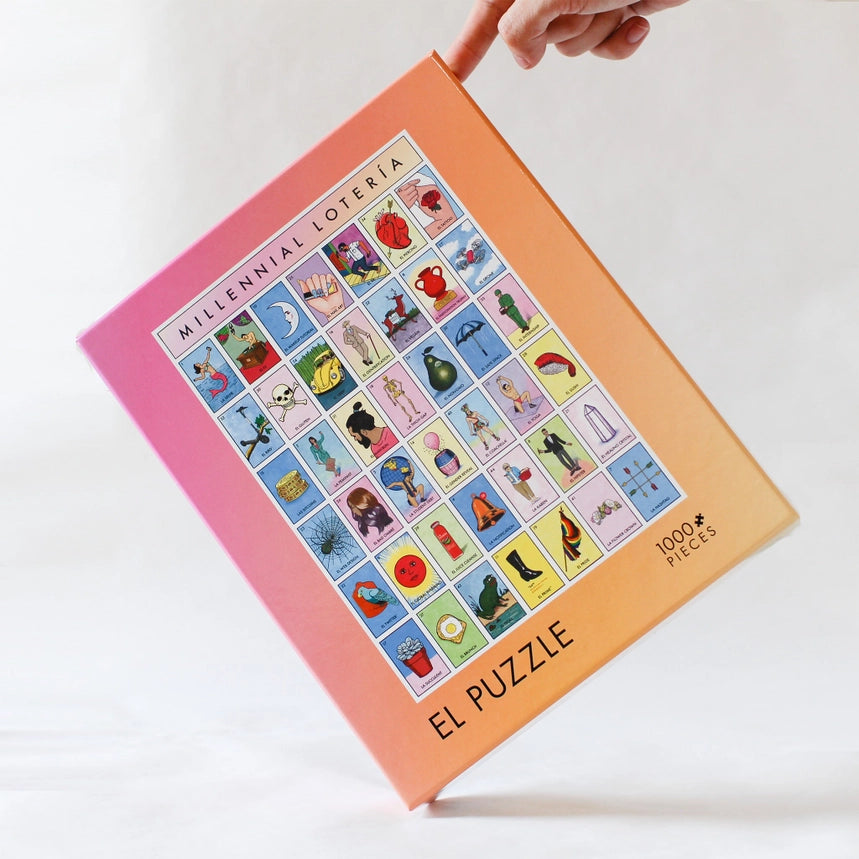 A hand holds a 1000-piece jigsaw puzzle box titled 'Millennial Loteria - El Puzzle'. The box features a colorful grid of various illustrated tiles on a peach-to-pink gradient background, set against a clean white studio backdrop.