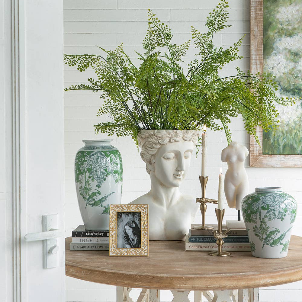 A styled interior scene showing a round wooden console table with a white carved pedestal base, holding a large green fern arrangement in a white classical bust vase, a green-and-white ceramic vase, a small framed print, books, and candlesticks. A white paneled wall and door frame surround the setup.