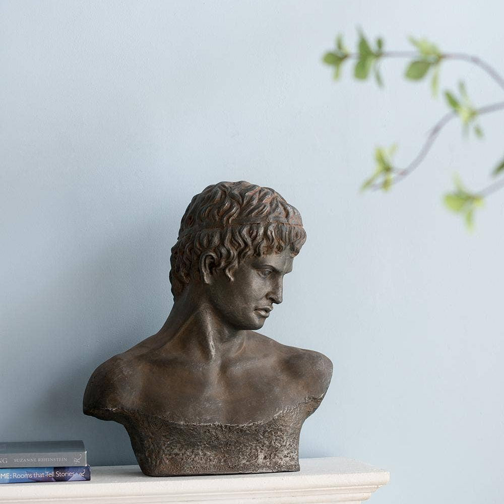 A classical-style bronze male bust sculpture displayed on a white mantelpiece against a pale blue wall, with a small stack of books at left and blurred green leaves in the upper right.