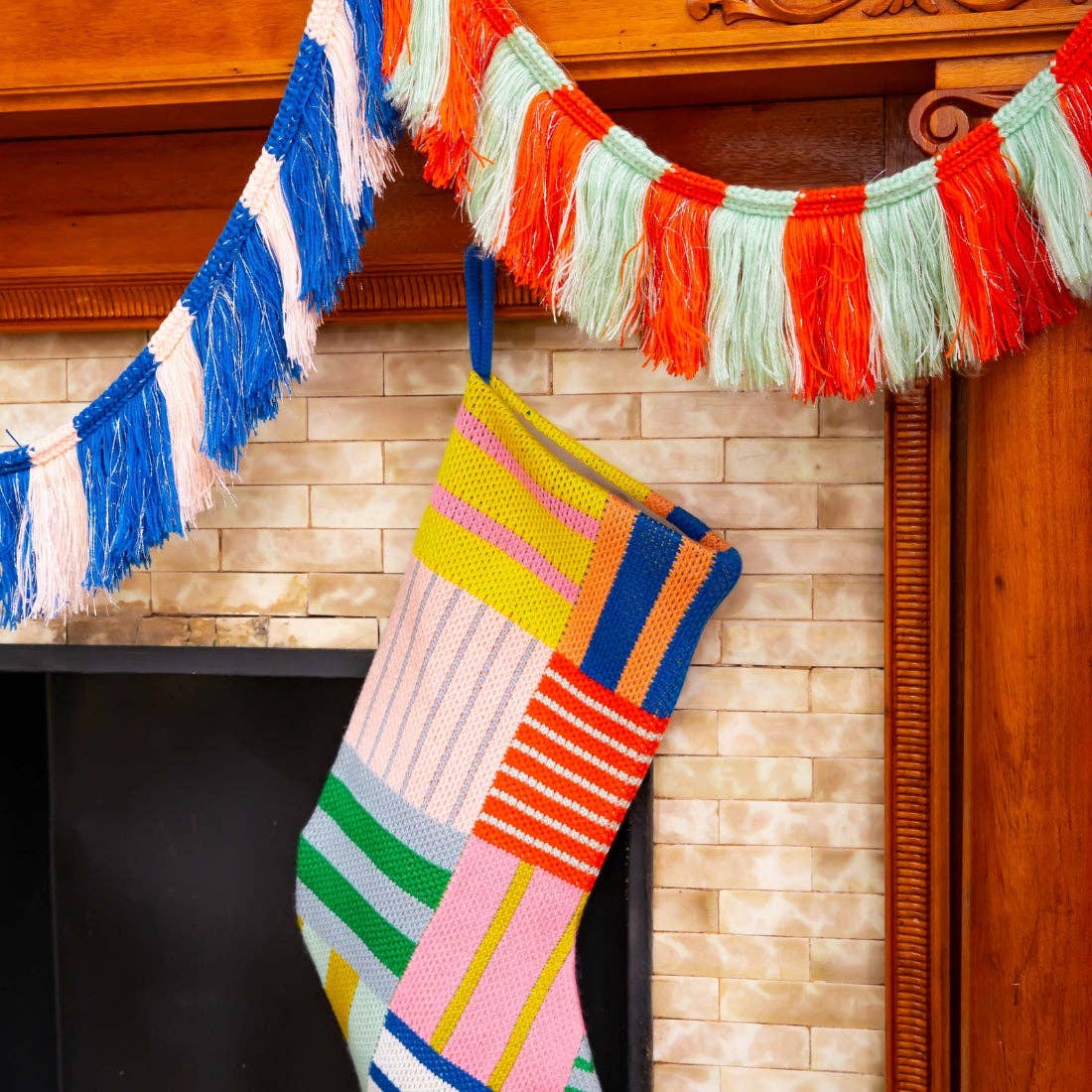 A colorful knit Christmas stocking hangs in front of a fireplace, decorated with striped and patchwork panels in pink, yellow, blue, green, orange, and white. A matching blue and white fringe garland and an orange, mint, and red fringe garland hang above the mantel.