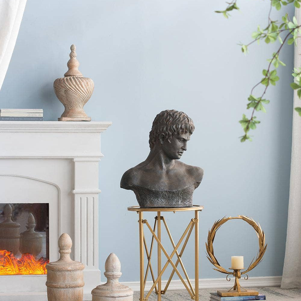 A dark bronze classical male bust sculpture displayed on a gold metal rolling stand in a styled room setting with a fireplace, decorative urns, and a light blue wall.