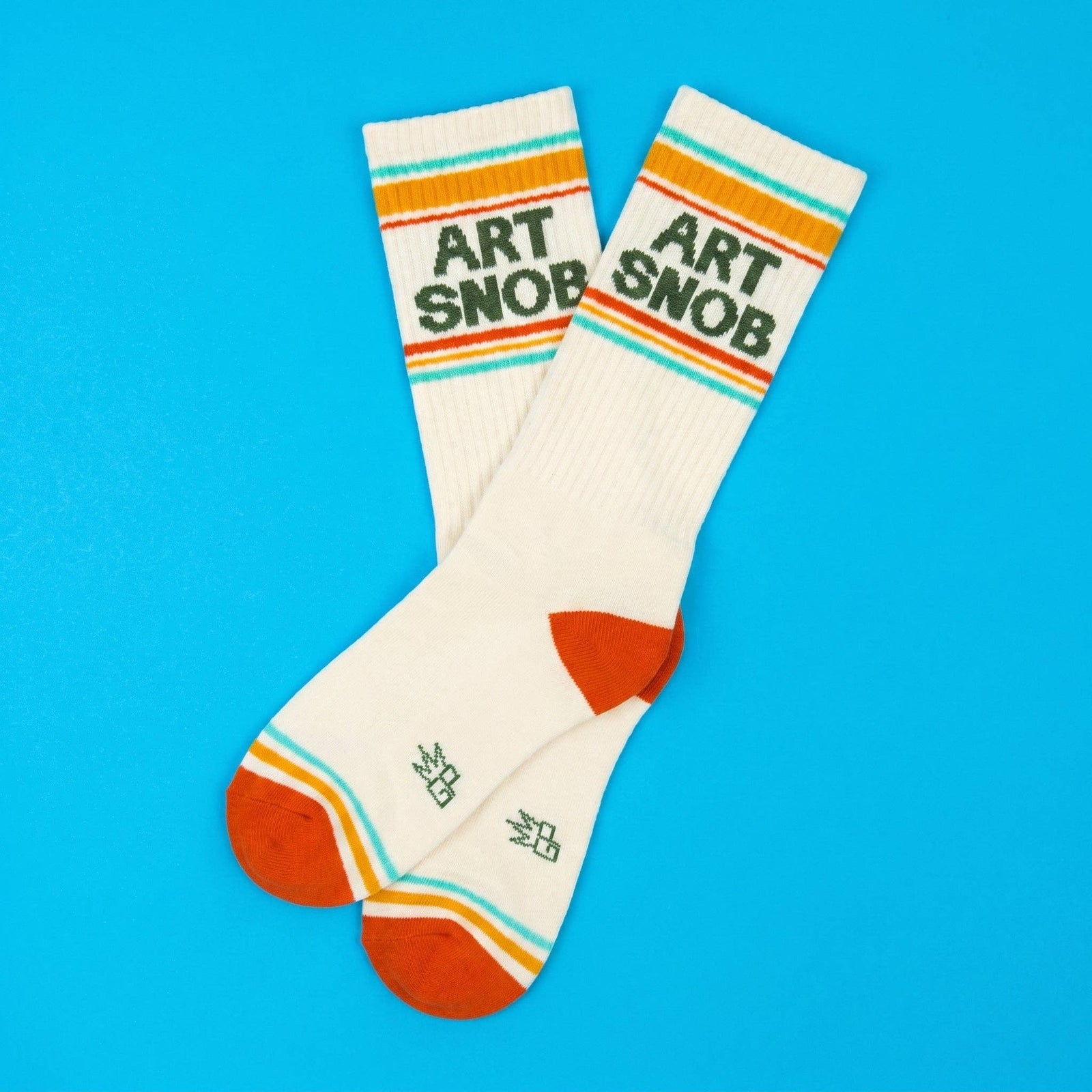 A pair of cream-colored crew socks with retro orange, yellow, and light blue stripes. The front sock features the text 'ART SNOB' in dark green.