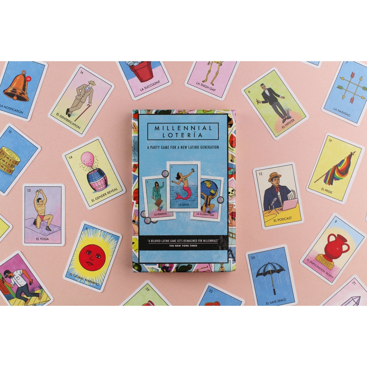 A boxed party game called 'Millennial Loteria' sits centered on a pink surface, surrounded by colorful illustrated playing cards spread across the scene.