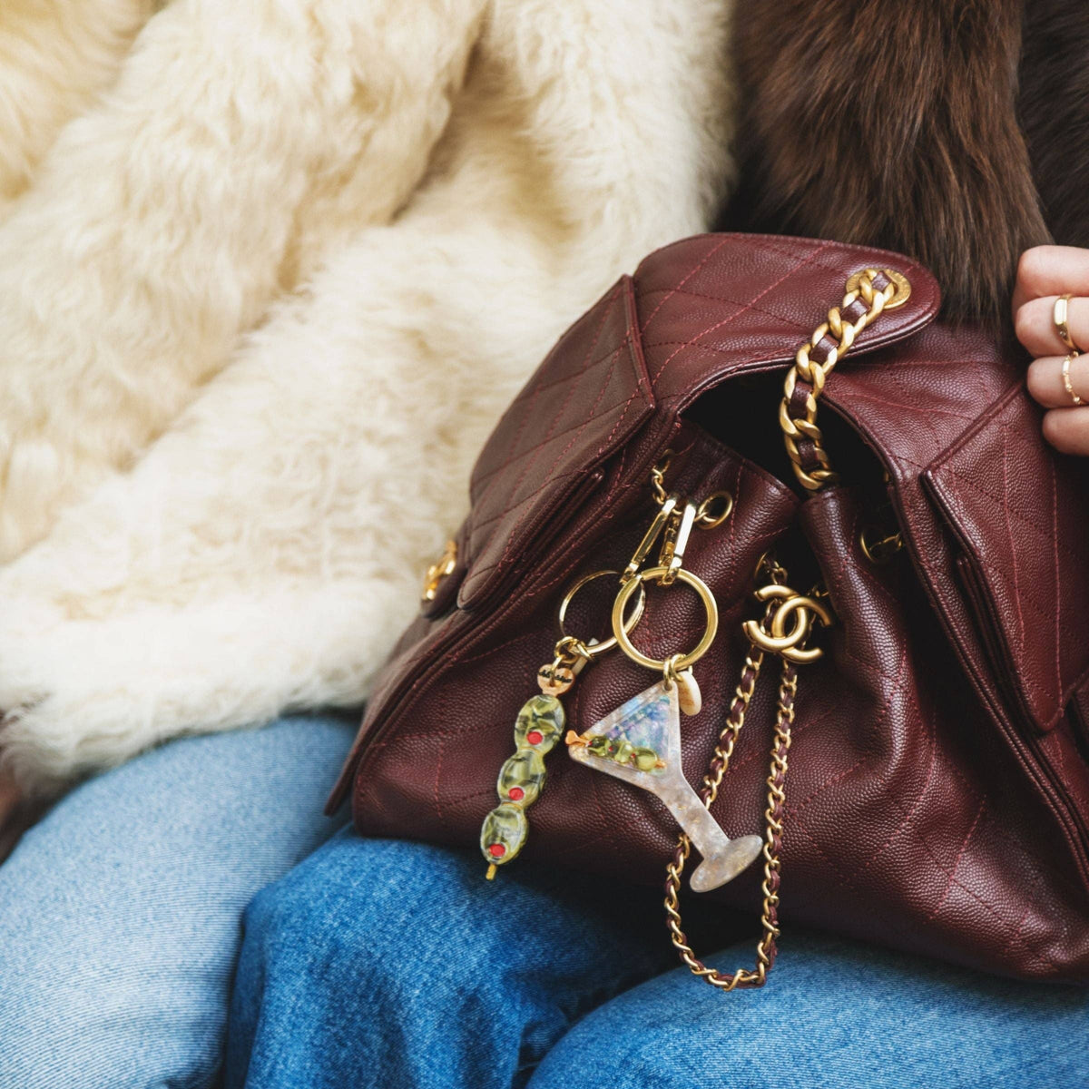 Brown leather handbag with gold chain and keychain on a person wearing blue jeans and a fur coat.
