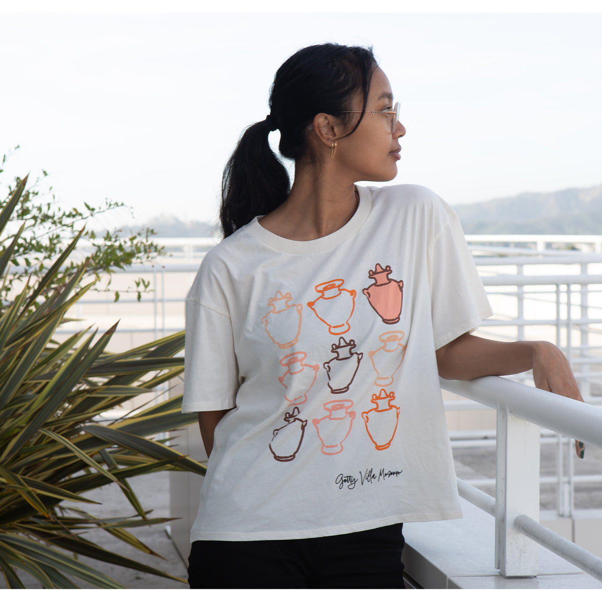 Woman wearing a white t-shirt with colorful graphic designs, standing on a balcony with plants and a scenic view.