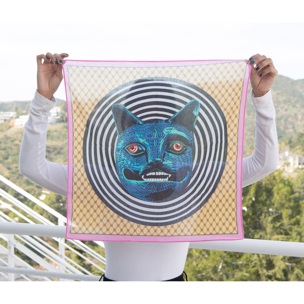 Person holding a square scarf with a blue cat design against a scenic background