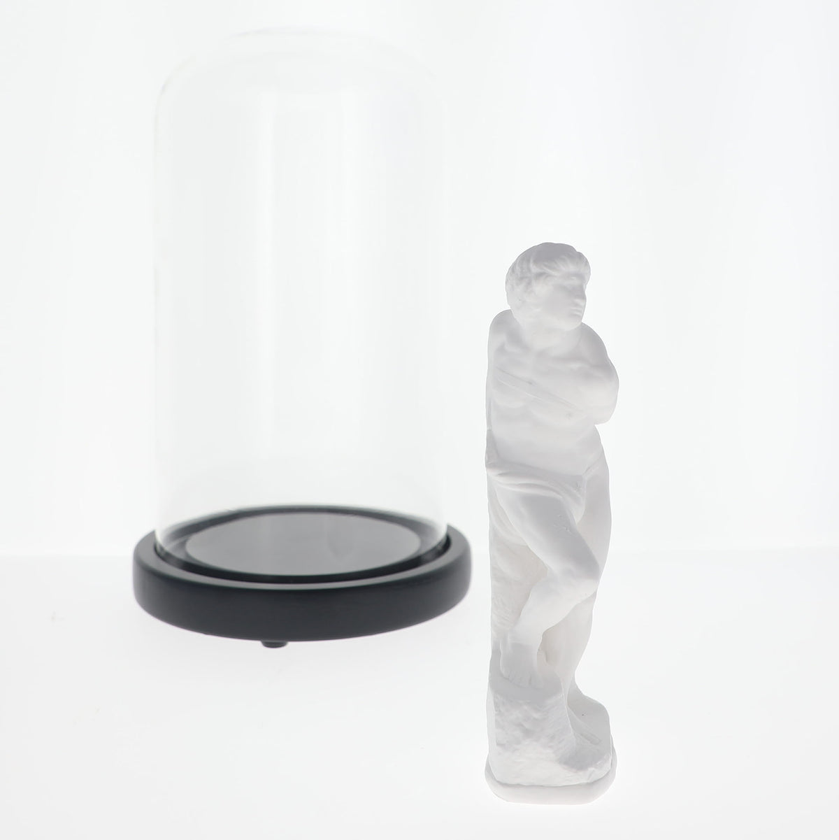 A frosted white classical-style human figure sculpture displayed next to a clear glass dome with a black circular base on a white studio background.